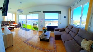 Ein geräumiges Wohnzimmer mit einer großen Couch, einem Couchtisch und einem an der Wand befestigten Fernseher. Es gibt einen Balkon mit Meerblick.