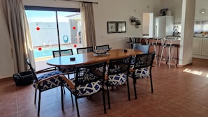 A dining area with a wooden table and chairs, a kitchen in the background, and a view of the sea through large windows. - Villa Georgiana Spacious Quiet 4 Bed Property With Private Pool Sleeps 8 (Yaiza)