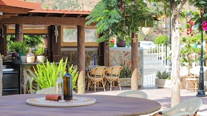 Une terrasse extérieure avec une table ronde et des chaises en osier, entourée de plantes en pot et d’un mur en pierre.