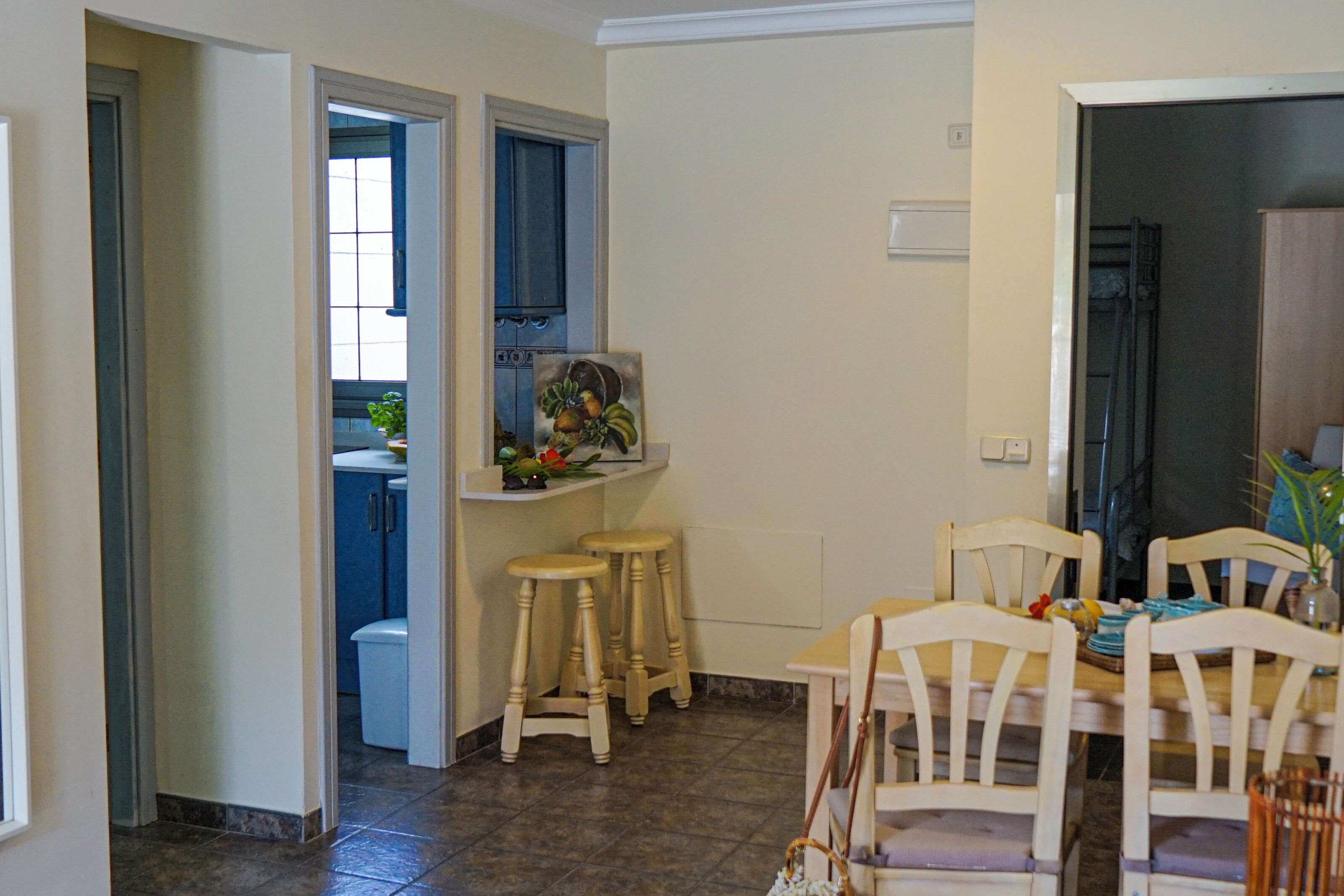 A dining area with a table and chairs, a painting on the wall, and a kitchen in the background.