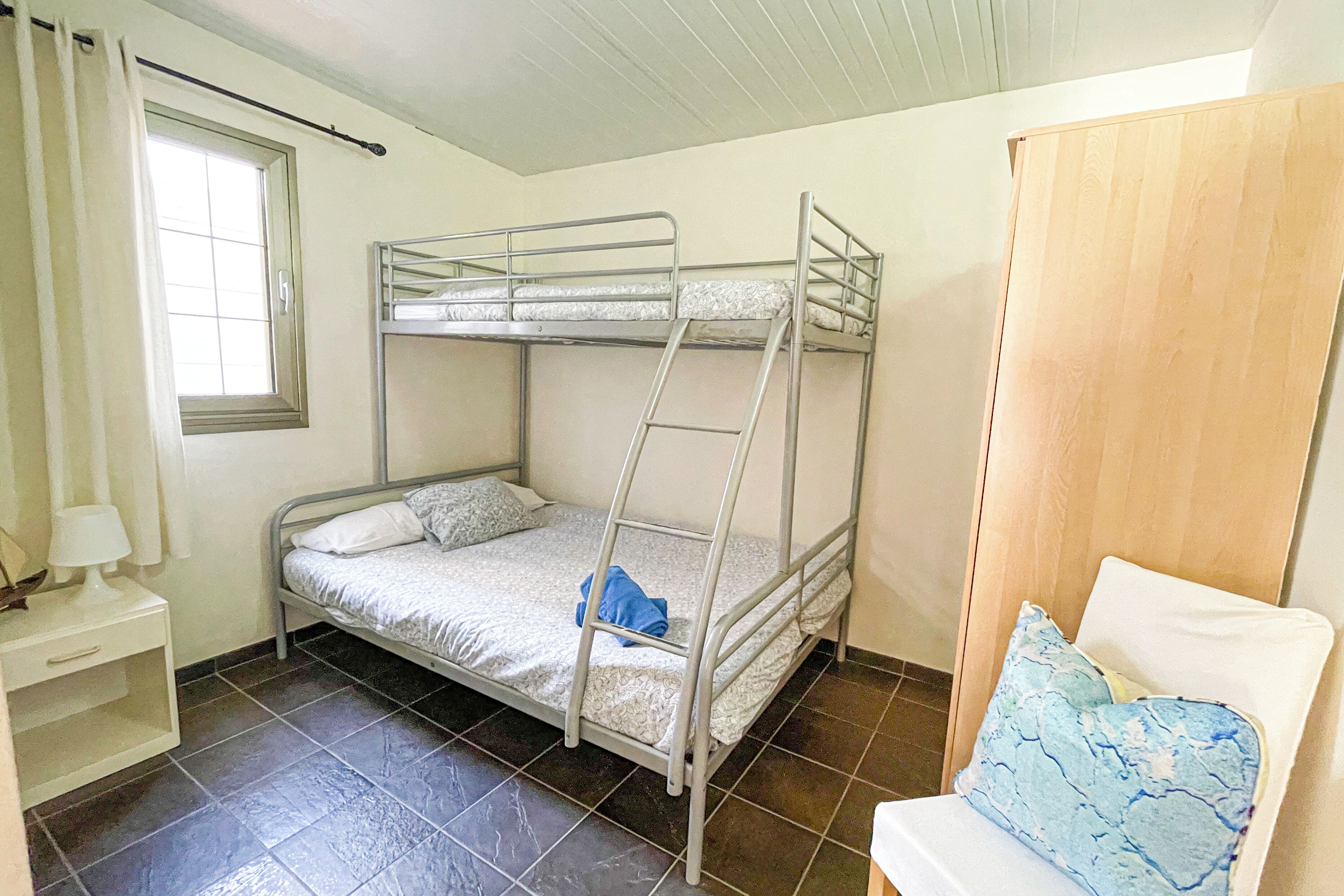 A bunk bed room with a window, a bedside table, and a chair with a cushion.