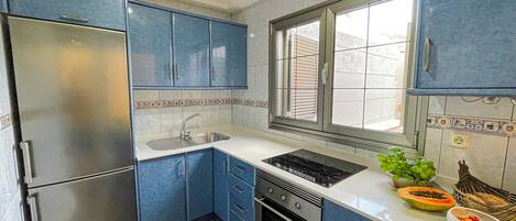 A compact kitchen with blue cabinets, a stainless steel refrigerator, a sink, and a window with blinds.
