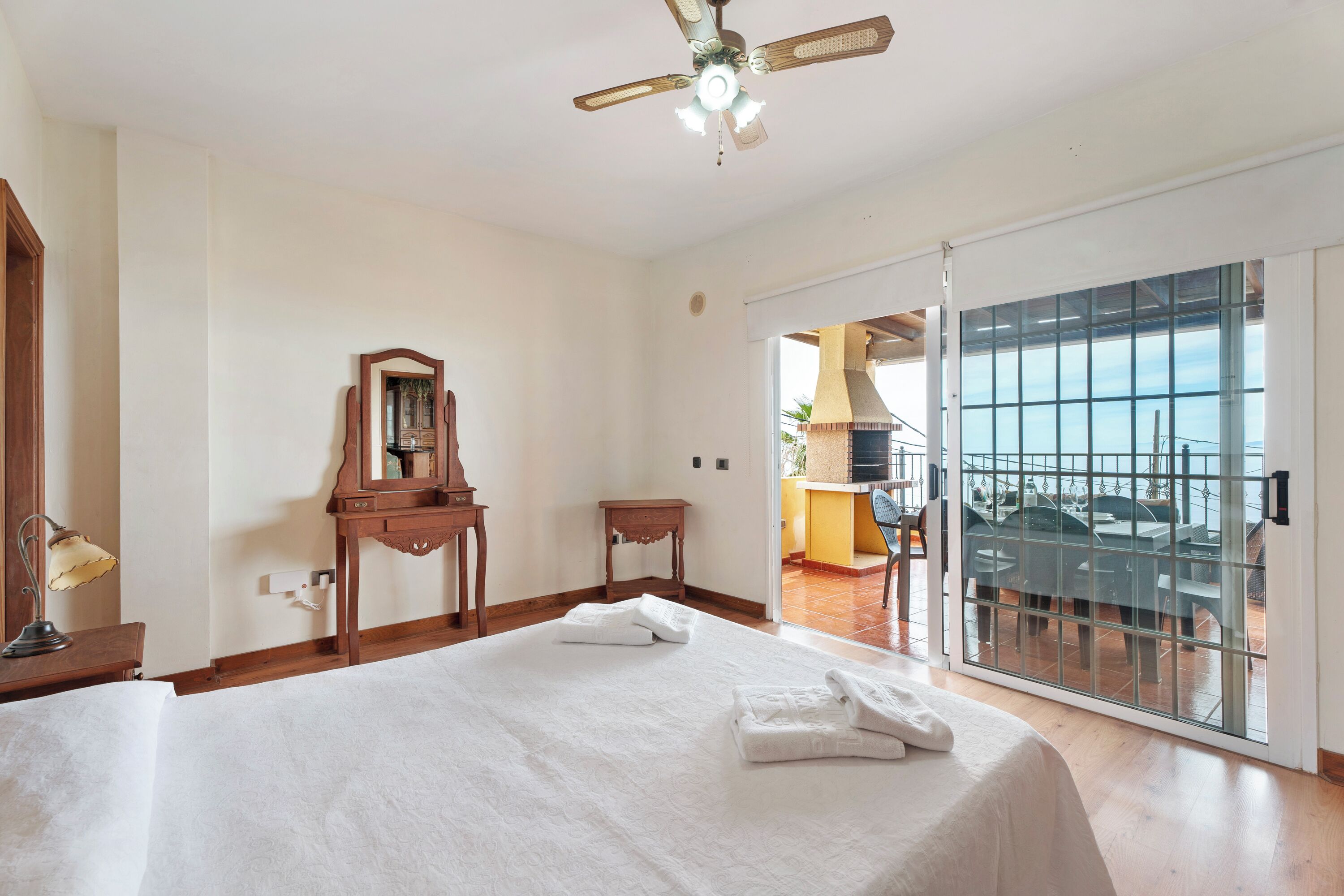 Un dormitorio con una cama grande, un ventilador de techo, un aparador de madera, un espejo y una puerta corrediza de vidrio que da a un balcón con mobiliario exterior.