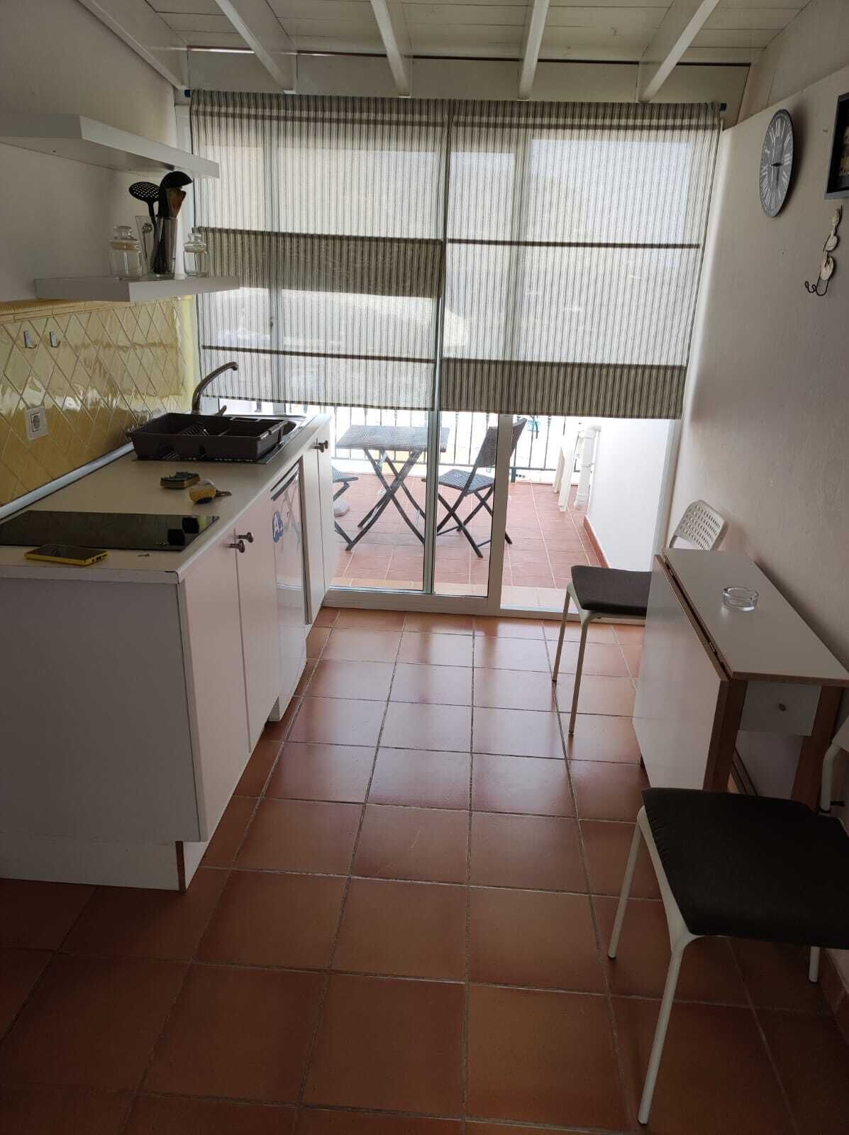 A compact kitchen with a sink, a small dining table, and a chair. There is a window with blinds and a view of the outside.