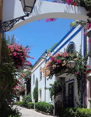 A narrow street with white buildings, arched doorways, and hanging flowers. - Charming 1 Bedroom Apartment in Puerto de Mogán (Mogan)