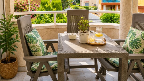 A covered outdoor seating area with wicker furniture, a table with a pitcher and glasses, and a view of a garden and buildings. - Beautiful And Bright Apartment In Calafell, (El Vendrell)