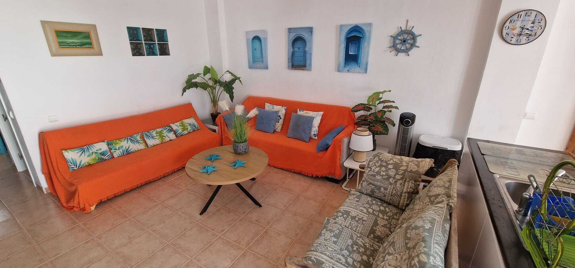 A living room with a sofa, a round coffee table, and a patterned sofa. There are wall-mounted artworks and a clock.