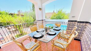 A balcony with a table set for four, surrounded by wicker chairs, overlooking a swimming pool and greenery. - Welcome to Paradise - Bienvenidos al Paraiso (Coin)