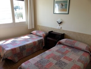 Ein Zimmer mit zwei Einzelbetten, einem kleinen Holzkommode, einem gerahmten Bild an der Wand und einem Fenster mit Vorhängen.