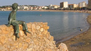 Eine Statue einer sitzenden Person auf einer Steinmauer an einem Ufer, im Hintergrund Gebäude.