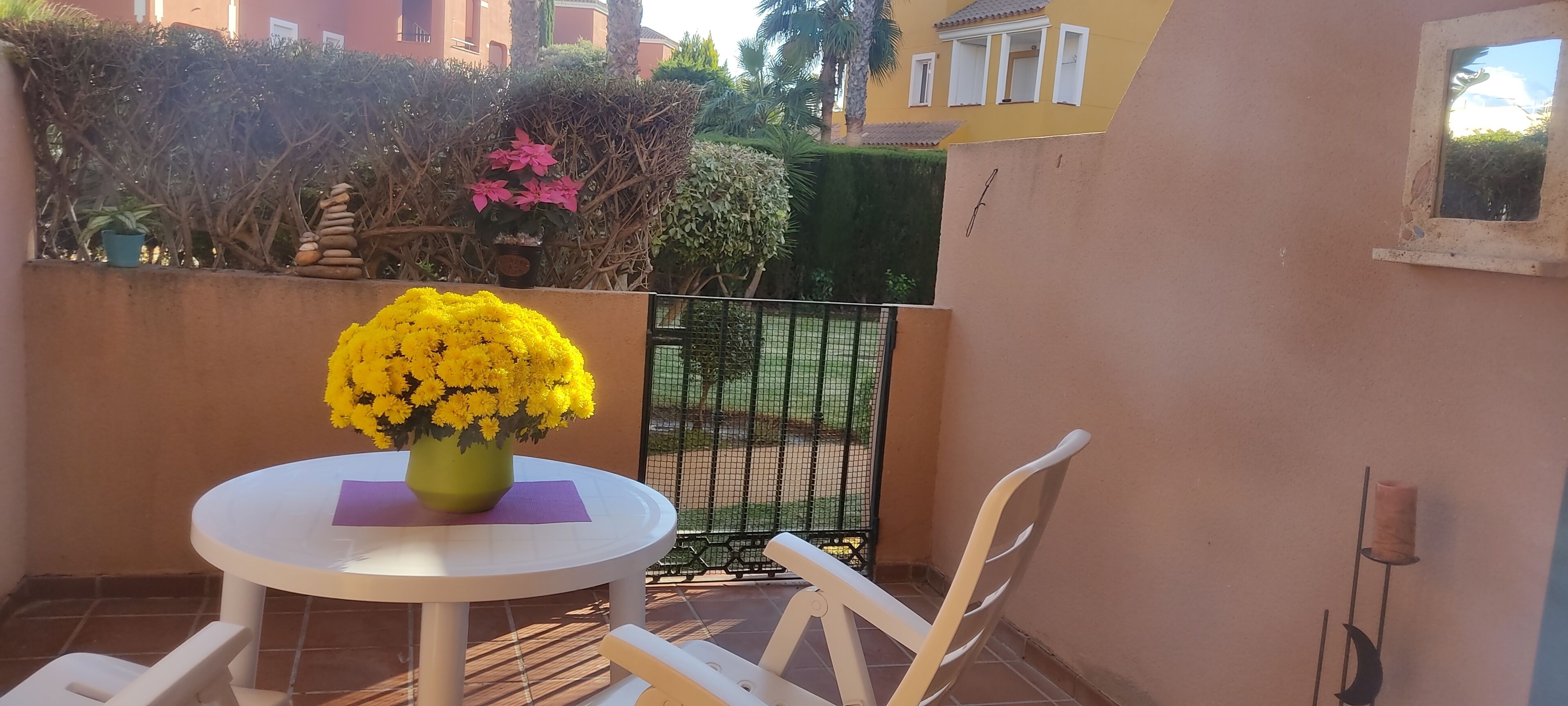 Un pequeño patio al aire libre con una mesa redonda y sillas blancas, una planta en maceta con flores amarillas, y una vista de un jardín y un edificio al fondo.