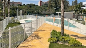 Zona de piscina con terraza cercada, palmeras y vista de edificios al fondo.