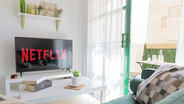 A modern living room with a flat-screen TV displaying 'NETFLIX', a white coffee table with a book and a cup, a green sofa, and a window with sheer curtains. - Casa Frida With High-speed Wifi and Smart-tv (La Oliva)