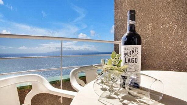 A table with a bottle of Puente del Lago wine, a glass, and a vase with flowers on a balcony overlooking the sea. - Costa Isora First Line and Pool (Guia de Isora)