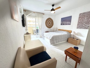 A hotel room with a bed, a desk, a chair, a ceiling fan, and a view of the outside.