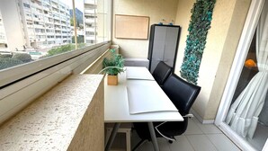 Un balcone con tavolo bianco, sedie nere, una pianta in vaso, un mobile grigio e un pannello decorativo.