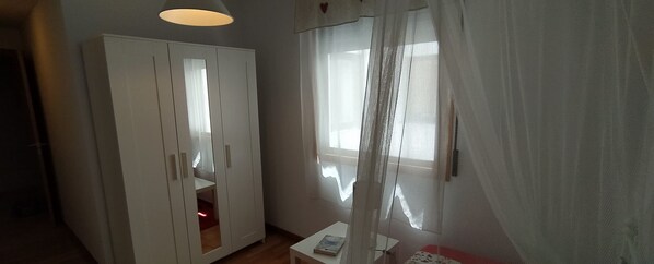 Une pièce avec une armoire, une petite table sur laquelle se trouve un livre, et une fenêtre ornée de rideaux légers.