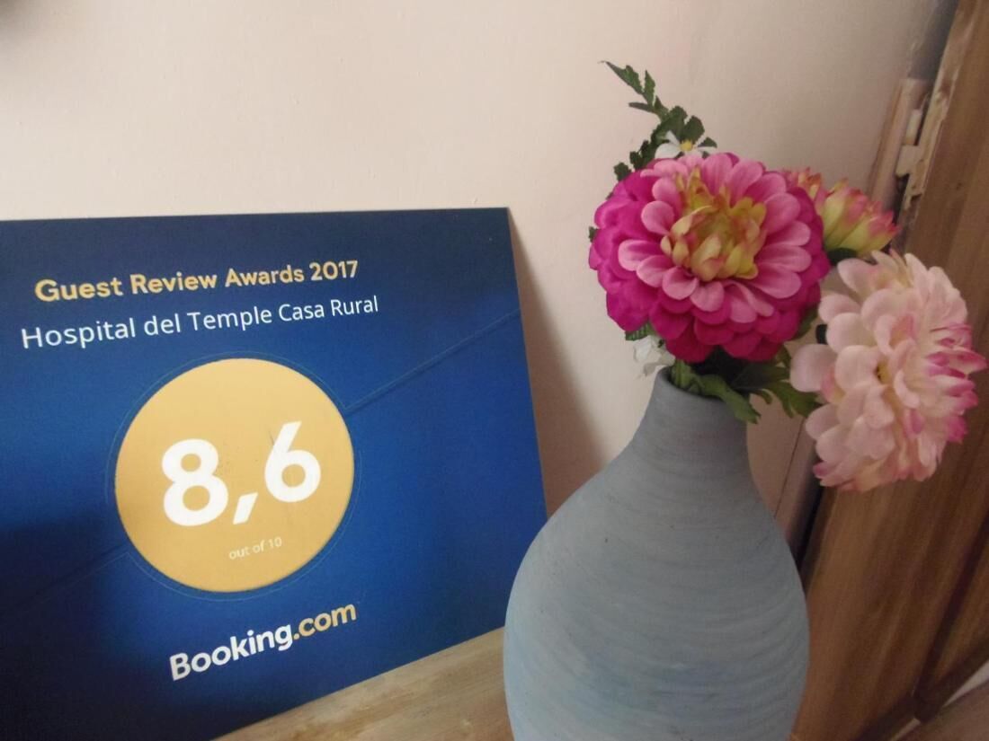 Señal azul con una calificación de 8.6 para el Hospital del Temple Casa Rural, logotipo de Booking.com y un jarrón con flores rosas.