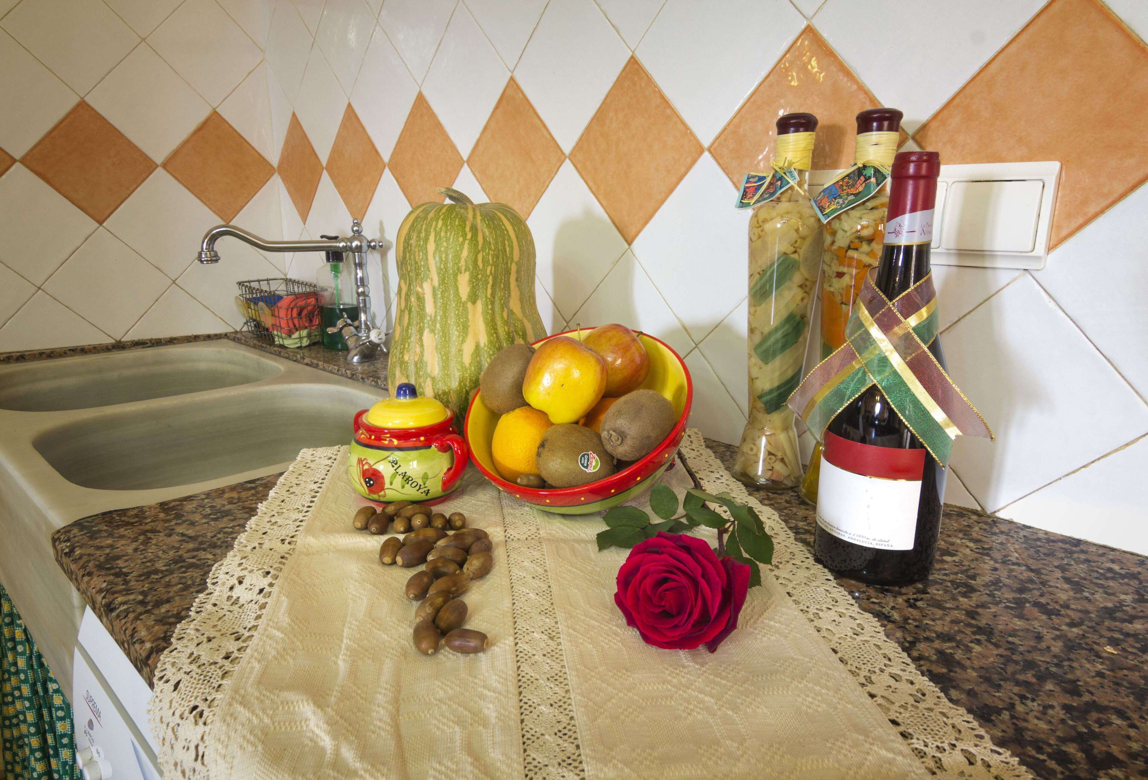 Una encimera de cocina con un lavabo, un cuenco de frutas, una botella de vino, una rosa roja y un objeto decorativo.