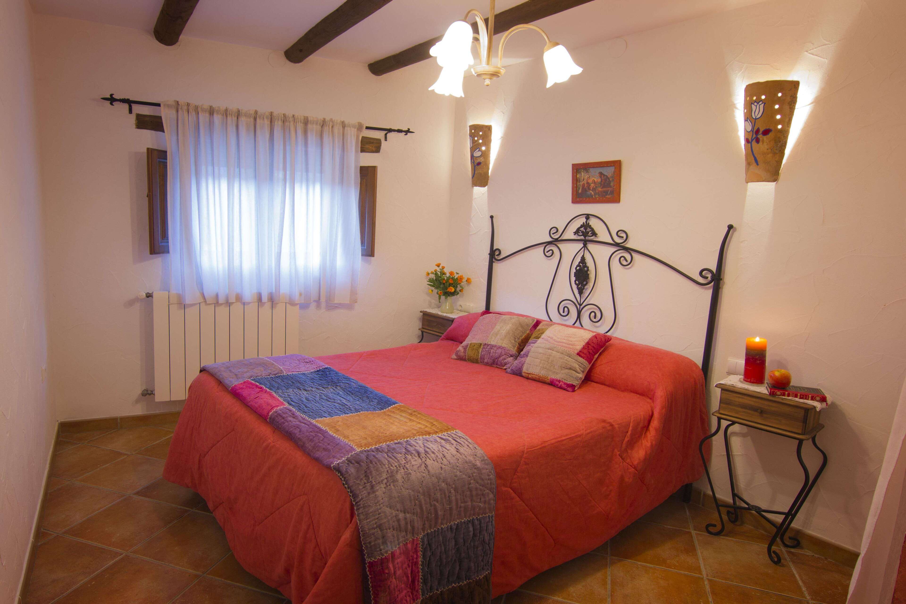 A bedroom with a red bedspread, a quilt, a bedside table with a candle, and a wall-mounted light fixture.