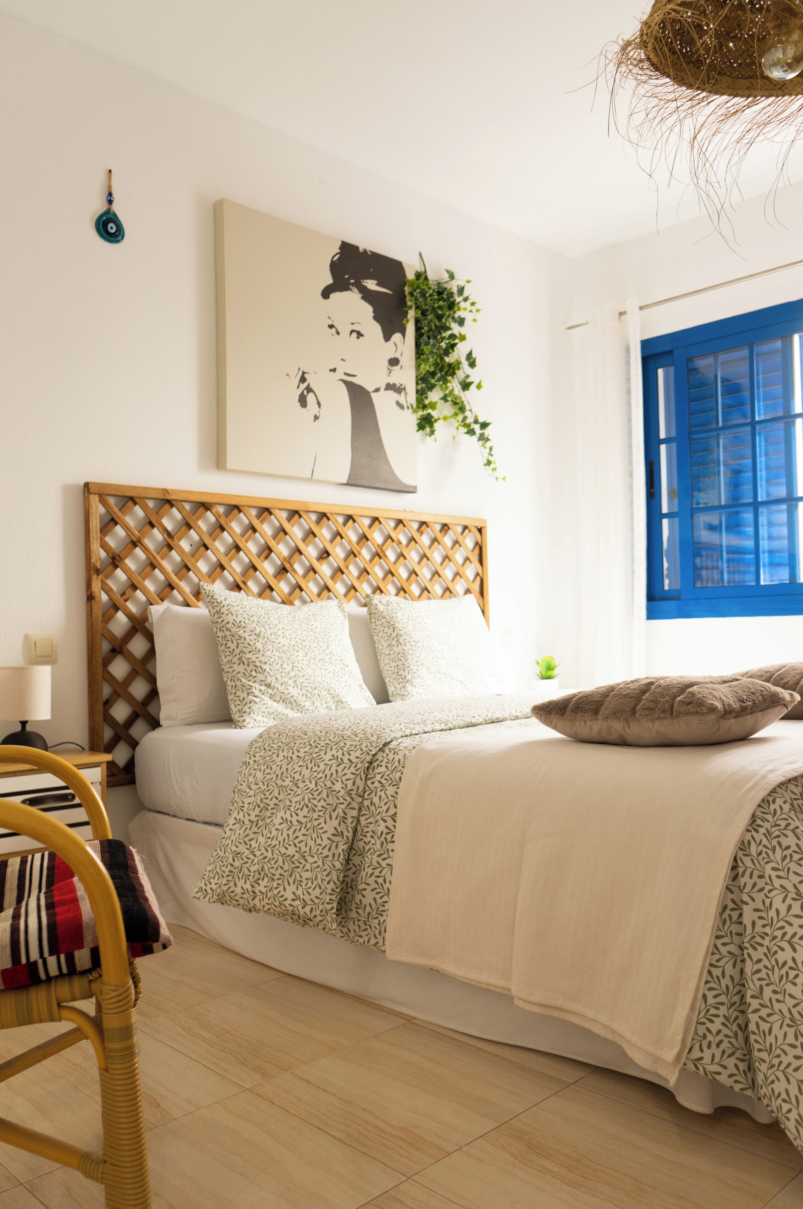 Un dormitorio con cabecera de madera celosada, una cama con ropa de cama estampada, una mesita de noche pequeña, una silla y una ventana con contraventanas azules.