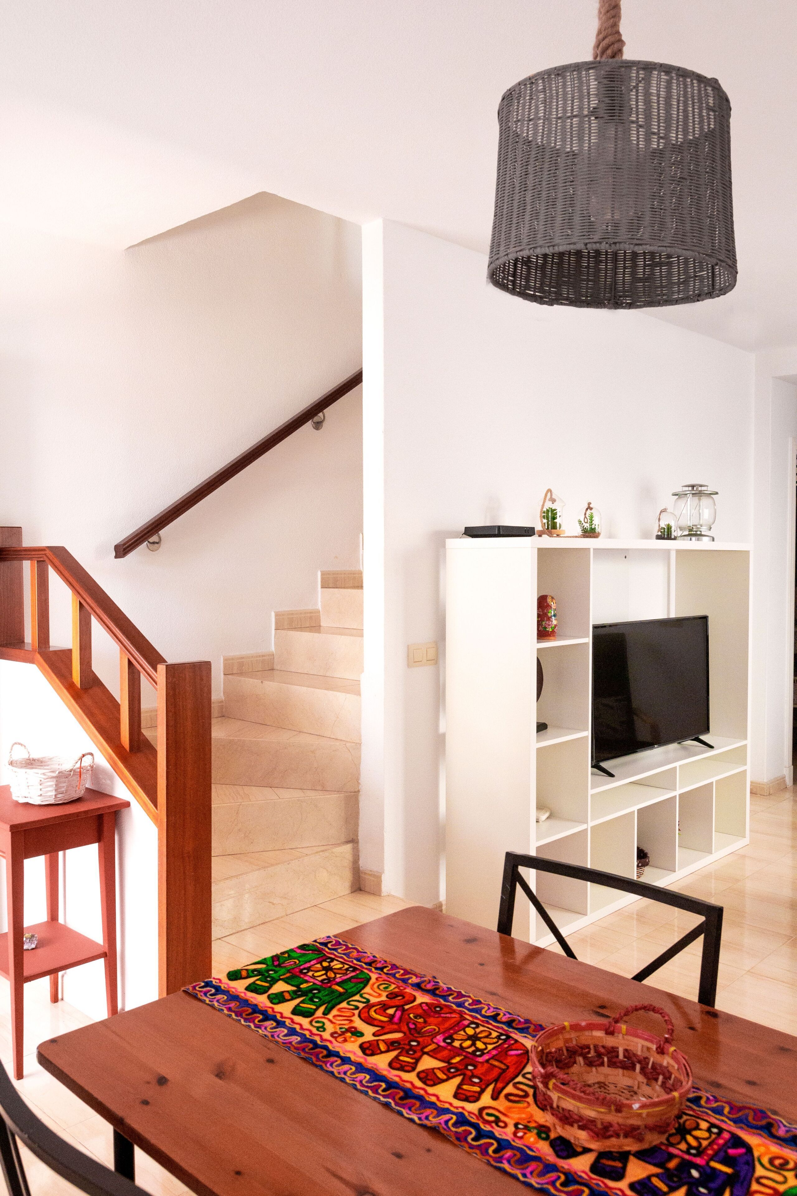 A modern living room with a wooden staircase, a white entertainment unit, and a dining table with a colorful placemat.