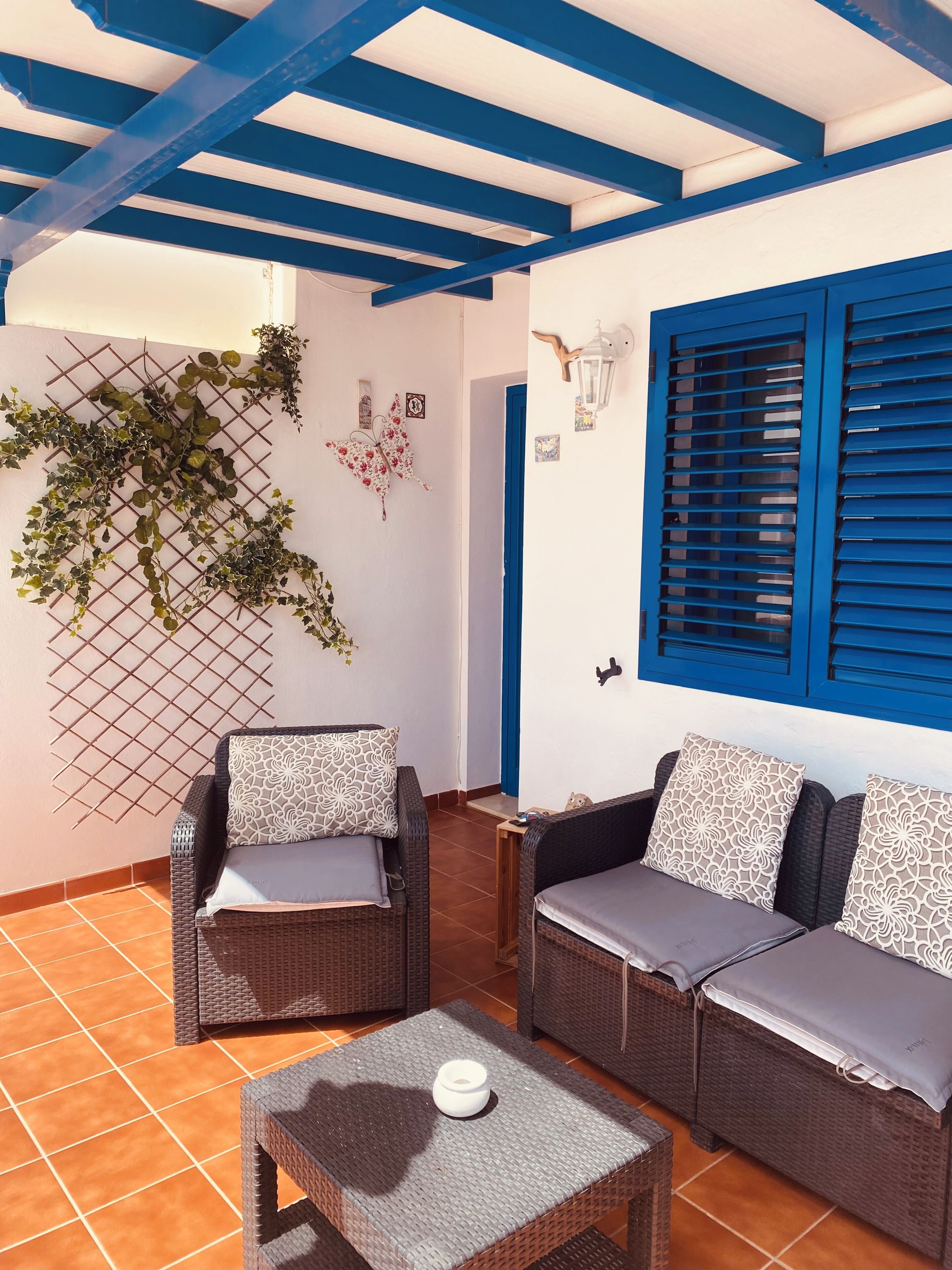 Un espace patio aménagé avec des meubles en osier, une petite table et un mur décoratif agrémenté de plantes et d’œuvres d’art mural.