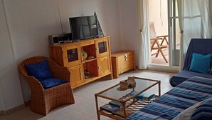 Ένα σαλόνι με έναν καναπέ, μια καρέκλα από μπαμπού, ένα τραπεζάκι σαλονιού και μια ξύλινη βιβλιοθήκη με μια τηλεόραση.