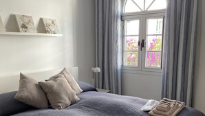 A neatly made bed with a blue quilt, pillows, and folded towels. There is a window with curtains, a radiator, and wall-mounted artwork. - Frontline Marina Apartment With Private Roofterrace and Seaview by El Sirocco (Mogan)