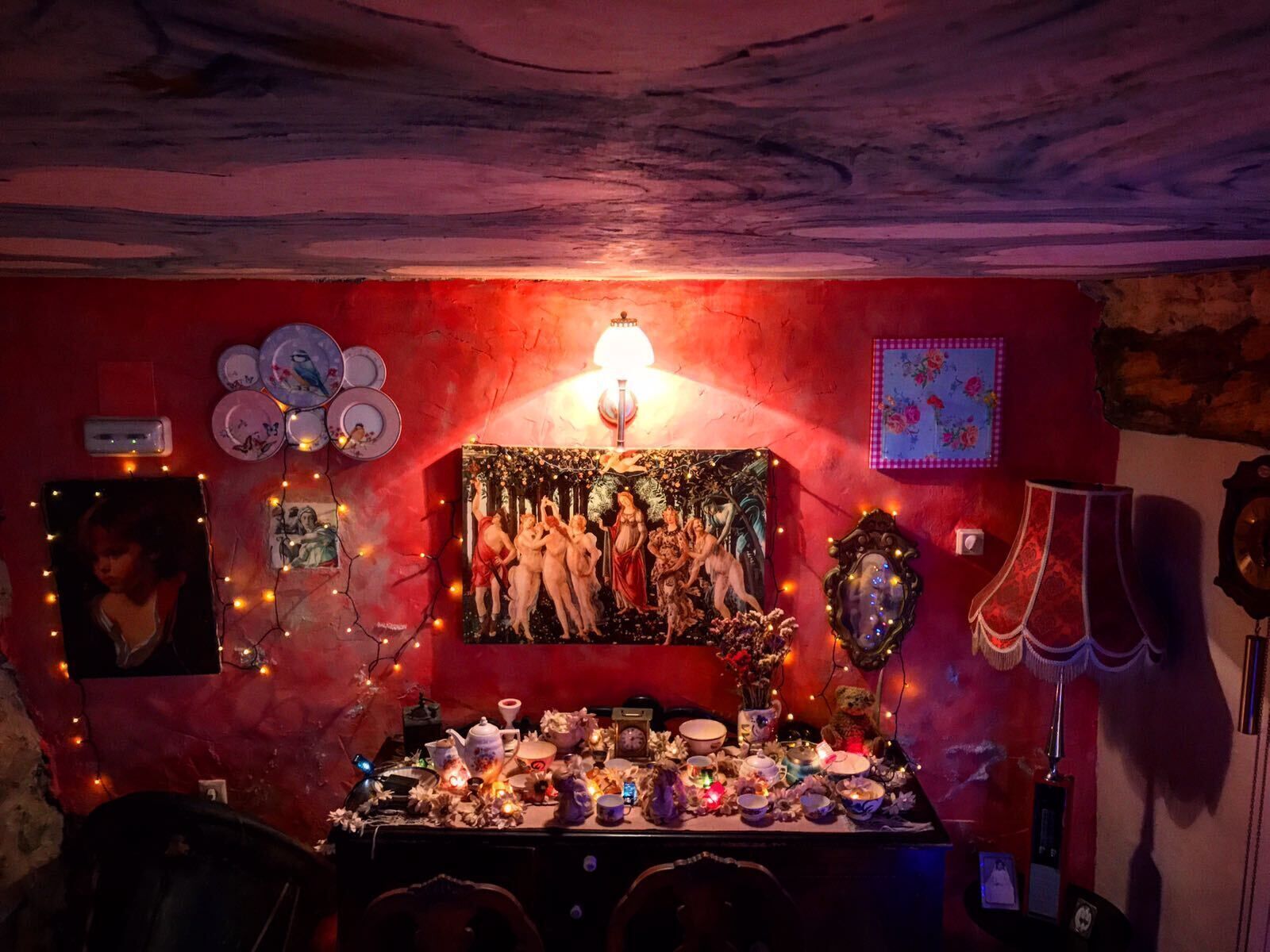 A room with a red wall, a painting of nudes, a table with various items, and string lights.