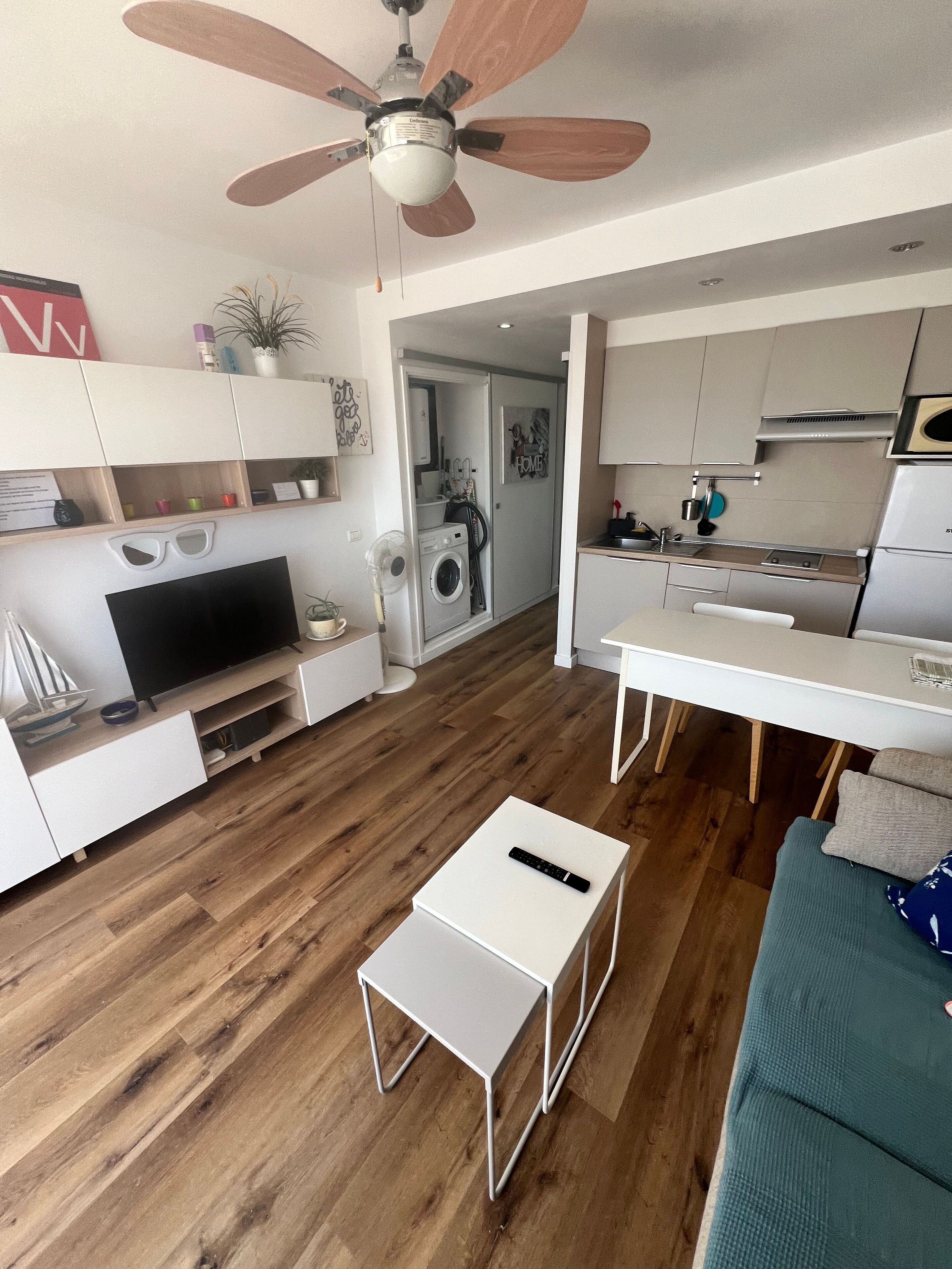 A modern living room with a flat-screen TV, a coffee table, a sofa, a ceiling fan, and a kitchen area with a refrigerator and stove.