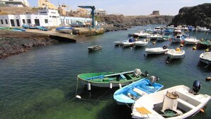 A marina with several small boats docked, a rocky shoreline, and buildings in the background. - Bravo´s Aparment Cotillo 1 (La Oliva)
