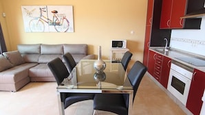 A modern living room with a glass dining table, black chairs, a grey sofa, and a red kitchen with white appliances.