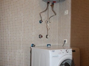 Una lavadora blanca con panel de control, un calentador de agua con tuberías y una pared con azulejos.