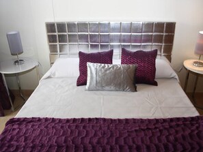 Una cama impecablemente arreglada con edredón blanco y dos almohadas moradas, además de una manta morada y un cabecero moderno.
