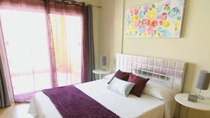 A neatly made bed with a white headboard, a purple throw blanket, and two pillows. There is a small bedside table with a lamp and a framed picture on the wall above the bed.