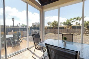 A glass-enclosed patio with a table and chairs, a small potted plant, and a view of a fenced outdoor area with trees and a street lamp. - Villa Marga, Villa At 100 Meters Far Of The Beach (La Oliva)