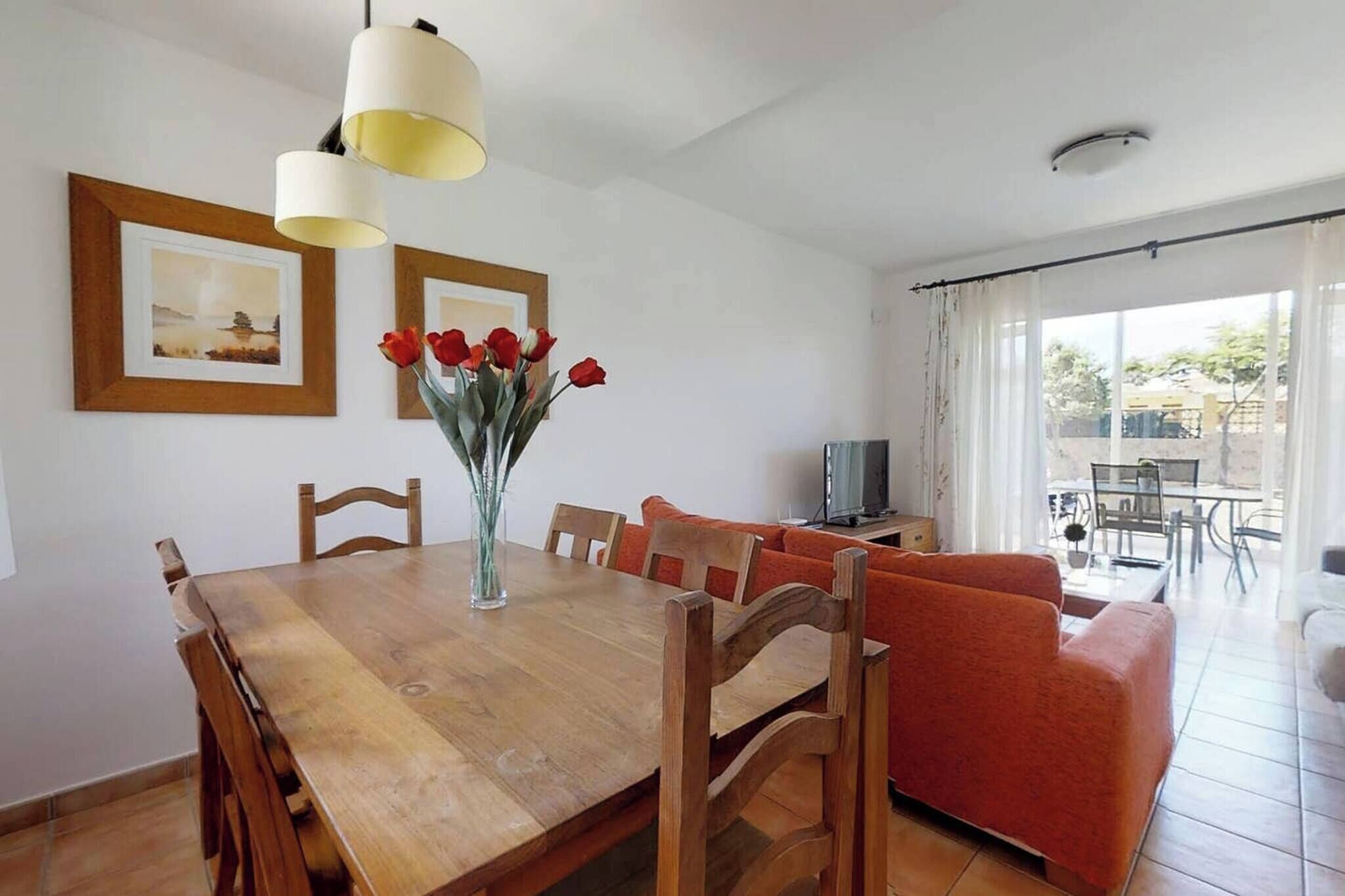 A dining area with a wooden table and chairs, a vase with red flowers, a sofa, and a television in the background.