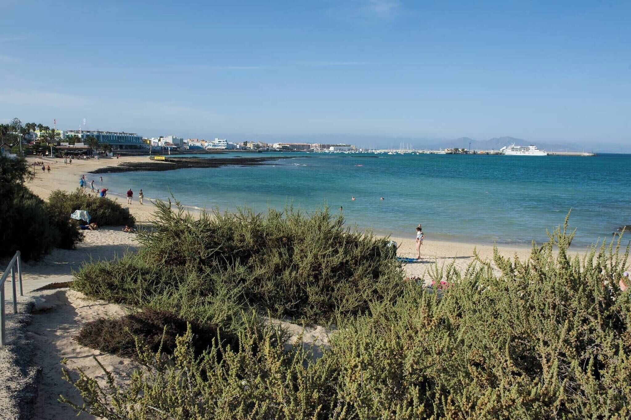 Une plage avec une eau d’un bleu limpide, un rivage sablonneux et quelques personnes. On aperçoit des bâtiments au loin et un bateau sur l’eau.