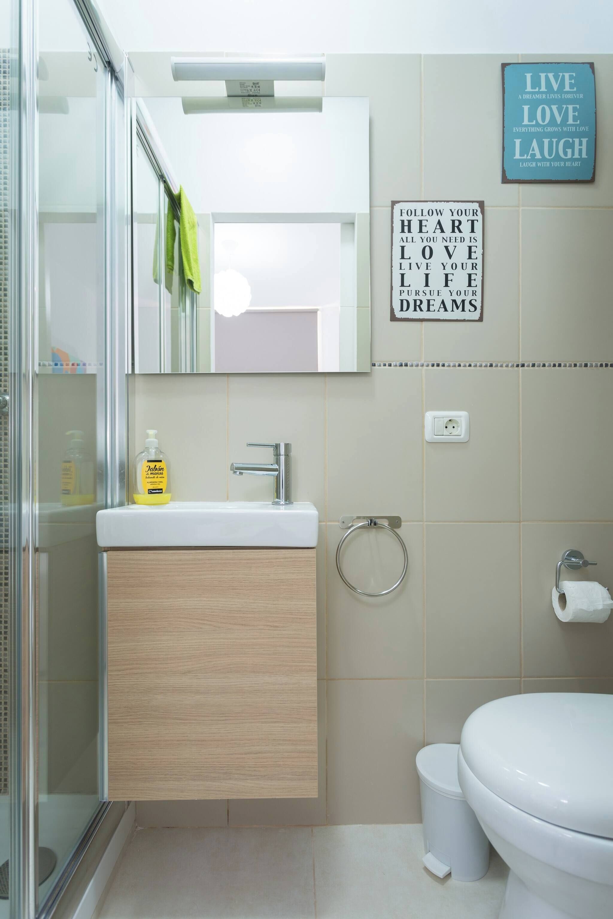 A modern bathroom with a glass shower enclosure, a wooden vanity, a white sink, and a toilet. There are motivational wall decorations and a towel hanging on the shower door.