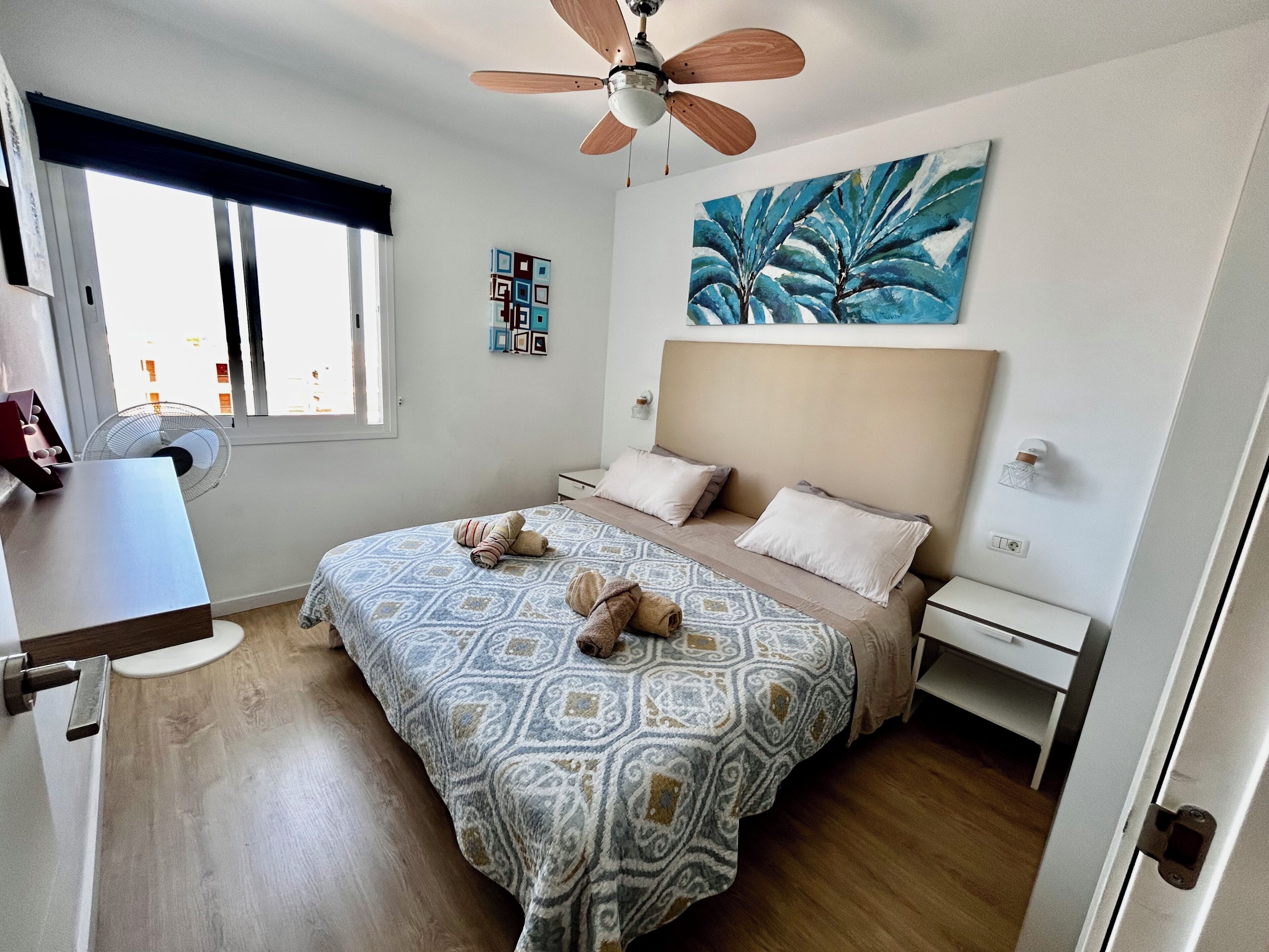 Une chambre à coucher avec un lit, des tables de chevet, un ventilateur de plafond, une fenêtre et un tableau.