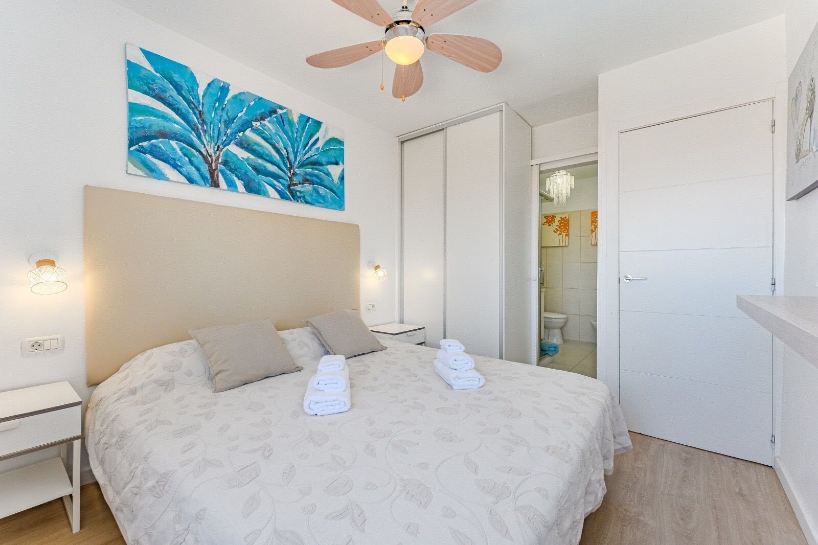 A hotel room with a bed, a ceiling fan, a painting, and a bathroom door.