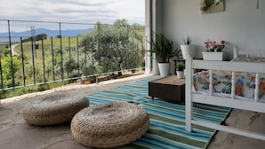 En balkon med udsigt over landskabet, en hvid seng, fletpuder og plantede planter.