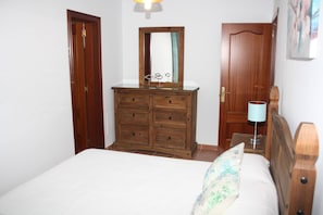 A bedroom with a wooden dresser, a bed with a floral pillow, a nightstand with a lamp, and a wooden door.