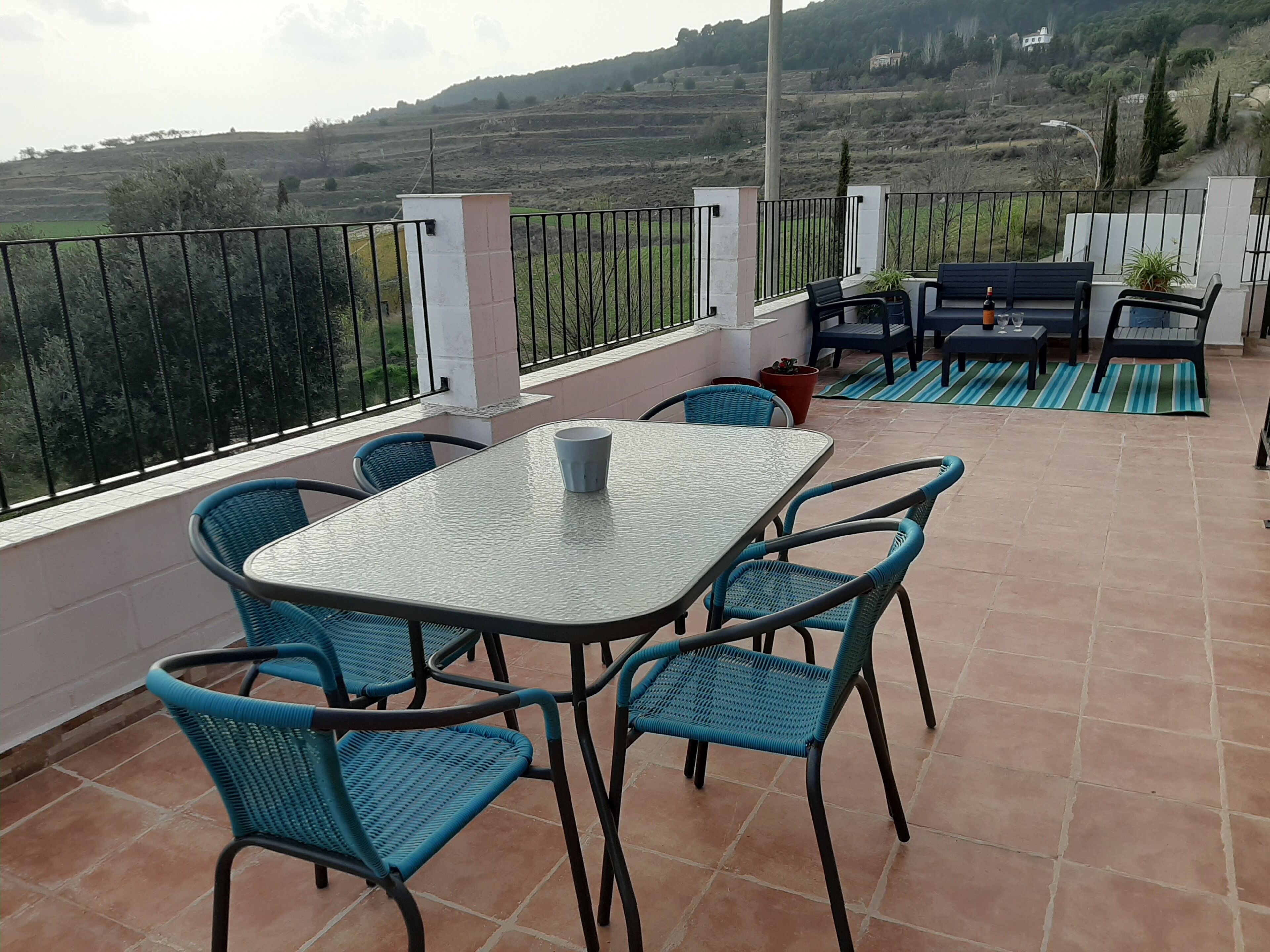Una terrassa amb taula i cadires, una taula petita amb una ampolla i un got, i vista d'un paisatge amb turons.