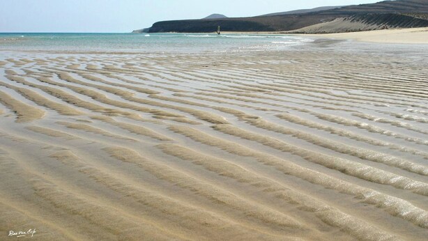 Una playa con patrones ondulados en la arena y una colina distante.