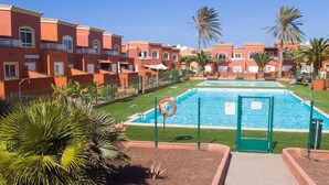 Un espace piscine avec une eau d’un bleu limpide, entouré d’une clôture, et situé à côté d’une rangée de bâtiments rouges dotés de balcons.