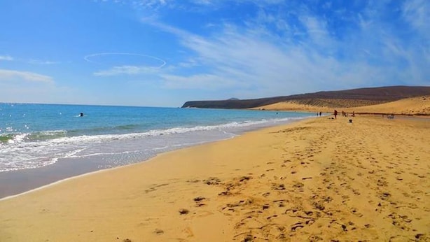 Une vaste plage de sable fin, parsemée d’empreintes qui mènent à l’eau, un ciel bleu azur et des collines lointaines.