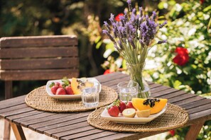 Uma mesa de madeira com um descanso de palha, um prato de frutas, um copo de água e um vaso com flores de lavanda.
