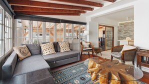 A modern living room with a gray sectional sofa, wooden beams, large windows, and a glass coffee table.